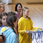 Dziewczynka w żółtym kombinezonie trzyma w rękach tacę z medalami. W tle grupa nauczycieli i uczniów.