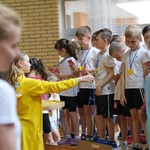 Dziewczynka w żółtym kombinezonie wręcza dzieciom medale.