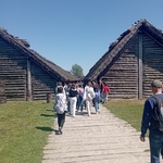 Grupa uczniów zwiedza stary gród słowiański w Biskupinie, w tle drewniane chaty.