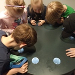 Dzieci pochylają się wokół okrągłego stołu i zaglądają w otwory ze szkłem powiększajacym.