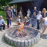 Grupa uczniów i rodziców piecze kiełbaski przy ognisku. W tle drewniana wiata.