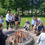 Uczniowie i rodzice stoją wokół ogniska i pieką kiełbaski.