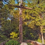 Miejsce pamięci oznaczone kamieniem i drewnianym krzyżem, w tle widoczne zielone drzewa.