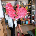 W bibliotece dwie uczennice trzymają czerwone papierowe serce. Zasłaniają nim swoje twarze..jpg