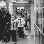 Wychowawczyni klas 1b prowadzi elegancko ubrane dzieci przez szkolny korytarz na uroczystość ślubowania.