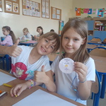 Dwie uśmiechnięte dziewczynki siedzą przy stoliku. Jedna z nich prezentuje otrzymany medal z okazji Dnia Dziewczynek..jpg