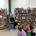 2 W bibliotece szkolnej starsi uczniowie czytają młodszym uczniom książkę. W tle półki z książkami i tablica informacyjna..jpg