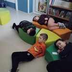 Grupa dzieci leżąca na pufkach w bibliotece szkolnej wsłuchuje się w słowa czytanej bajki.jpg