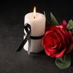 Biała świeczka z zawiązaną czarną wstążką i czerwona róża na czarnym tle.